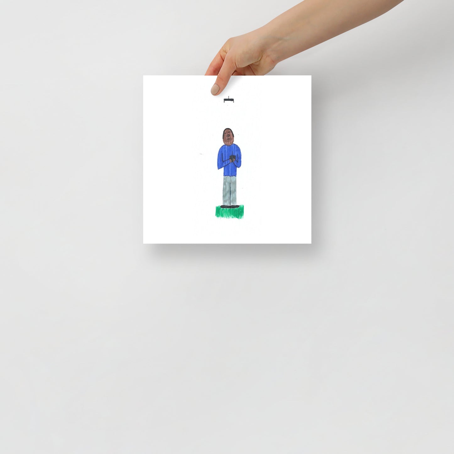This image shows an art print of a square illustration featuring a figure in a blue outfit standing on green ground flying a drone against a white background. Minimalist and simple design.
